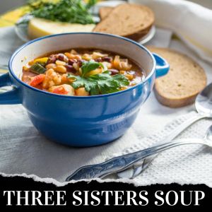 A blue bowl of Three Sisters Soup garnished with herbs. Two slices of brown bread, a yellow vegetable, and herbs in the background on a white cloth. Two spoons are beside the bowl.