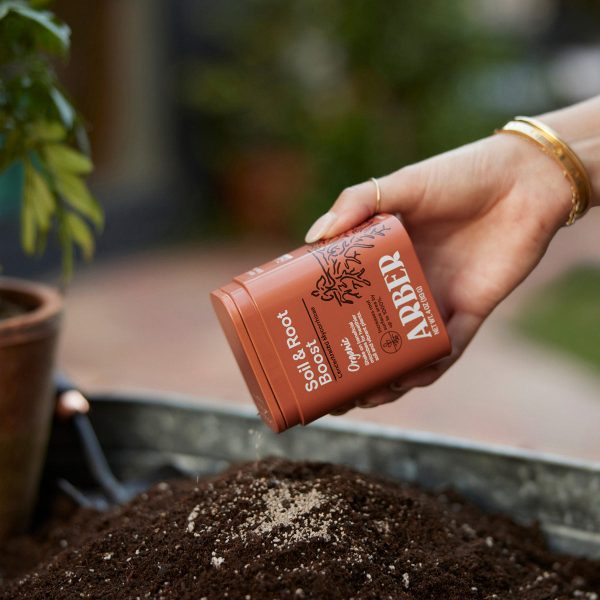 A hand sprinkles Arber Salt & Root Boost powder onto soil in a garden setting.