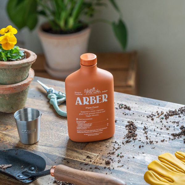 A bottle of Arber plant food sits on a wooden table with gardening tools, gloves, soil, small potted flowers, and a plant in the background.